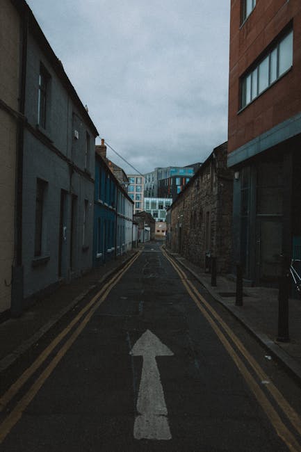 A narrow urban street with a single white arrow painted on the asphalt indicating the direction of travel. On the left side, there are low-rise buildings with dark-colored facades, some with small windows and doors. On the right side, a taller modern building with large glass windows and a reddish-brown exterior is visible. The street is lined with black bollards and a few pavement posts, with minimal street furniture. Overhead, power lines run across the sky, which is overcast with grey clouds, suggesting cloudy weather. This scene captures a typical narrow street that could be part of a home relocation or moving process, emphasizing confined spaces often encountered during furniture transport in urban environments managed by removal companies like Removal Companies Clapham.