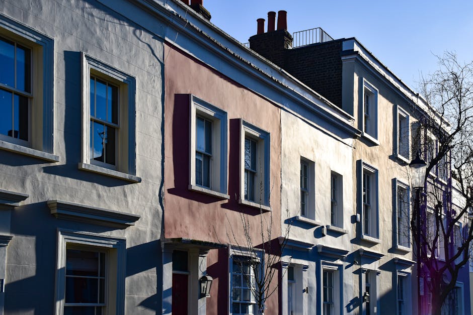 Photograph of the exterior of a residential building in Clapham Old Town, showing a row of terraced houses with a mix of white and pastel pink façades. The buildings feature multiple sash windows with white frames and decorative lintels. Some windows have shades or curtains visible through the glass. The image captures a clear daytime scene with sunlight casting shadows on the front of the houses, highlighting their architectural details. There are rooftop chimneys visible against a blue sky, and a leafless tree is present on the right side of the frame, indicating a winter or early spring season. This setting depicts a typical London-style neighborhood, where [COMPANY_NAME] might assist with the furniture transport, home relocation, or packing and moving services as part of a comprehensive removals process. The scene suggests a calm, residential environment suitable for loading and unloading furniture and boxes in preparation for a move or house clearance.