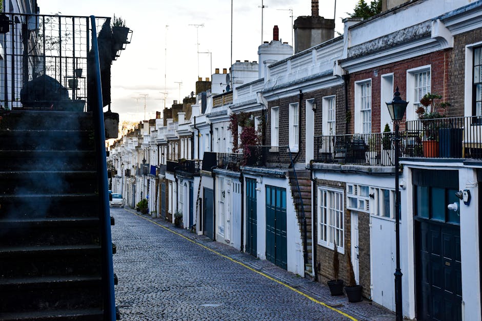 A row of terraced houses along a narrow cobbled street in Clapham Old Town during daylight, showing residential exteriors with brick and white-painted facades, small balconies with black wrought iron railings, potted plants, and traditional street lamps. A black metal staircase on the left side leads up to one of the houses, and parked cars are visible further down the street. The scene captures a typical residential area where house removals and furniture transport may take place, with No visible moving activity or equipment, but the setting suggests an environment suitable for home relocation services. Removal Companies Clapham offers assistance with packing and moving logistics in this area.