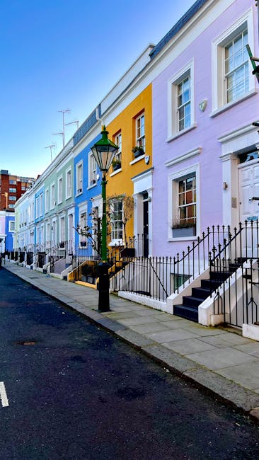 Photograph of the exterior of a residential building in Clapham Old Town, showing a row of terraced houses with a mix of white and pastel pink façades. The buildings feature multiple sash windows with white frames and decorative lintels. Some windows have shades or curtains visible through the glass. The image captures a clear daytime scene with sunlight casting shadows on the front of the houses, highlighting their architectural details. There are rooftop chimneys visible against a blue sky, and a leafless tree is present on the right side of the frame, indicating a winter or early spring season. This setting depicts a typical London-style neighborhood, where [COMPANY_NAME] might assist with the furniture transport, home relocation, or packing and moving services as part of a comprehensive removals process. The scene suggests a calm, residential environment suitable for loading and unloading furniture and boxes in preparation for a move or house clearance.