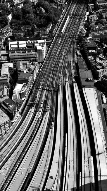 Black and white aerial photograph of a cityscape featuring multiple train tracks and railway lines that curve and intersect, running parallel to adjacent urban buildings and roads. The image captures the complex rail infrastructure in a dense urban area, with surrounding structures including modern office buildings, smaller residential and commercial properties, and greenery visible in the background. This scene illustrates the transportation and logistics environment relevant to home relocation and furniture transport services. Such detailed views of railway networks underscore the logistics involved in moving or delivering household items during professional removals by companies like Removal Companies Clapham, emphasizing the importance of efficient planning and transportation in house moving processes.
