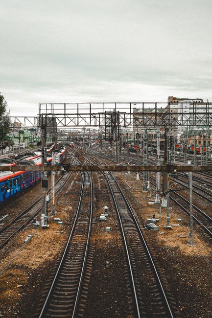 A wide view of multiple railway tracks and overhead electric wires extending into the distance, with several trains parked along the sides of the tracks. The scene includes metal signals and infrastructure supporting rail transport, set against an overcast sky. The tracks are situated on gravel beds, and a pedestrian bridge or similar structure can be seen crossing above the tracks in the background. This transportation environment is indicative of a busy station or rail yard. Although the image is primarily focused on railway infrastructure, it does not depict house removals or moving services directly. However, the detailed railway setting aligns with transportation and logistics themes relevant to furniture transport and home relocation processes, as part of comprehensive moving solutions supported by companies like Removal Companies Clapham.